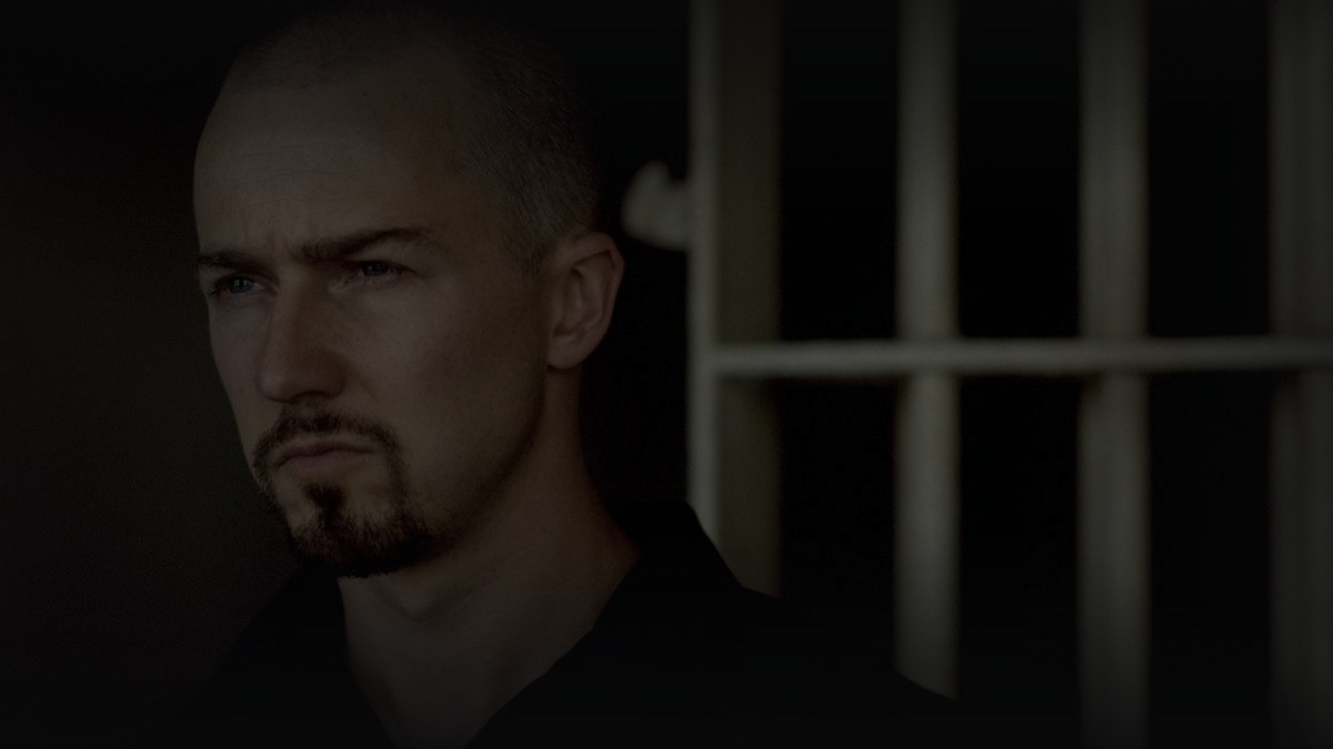 Still from American History X (1998) — 16:9 still from Letterboxd (the 1200x675 crop variant), baked through scripts/bake_dark_photo.py at default factor, mean brightness 22.3 (in canon range, on the dim side for the black-and-white flashback aesthetic of the source still). source: edward norton as derek vinyard, in prison, in front of bars - the canonical still for the film.